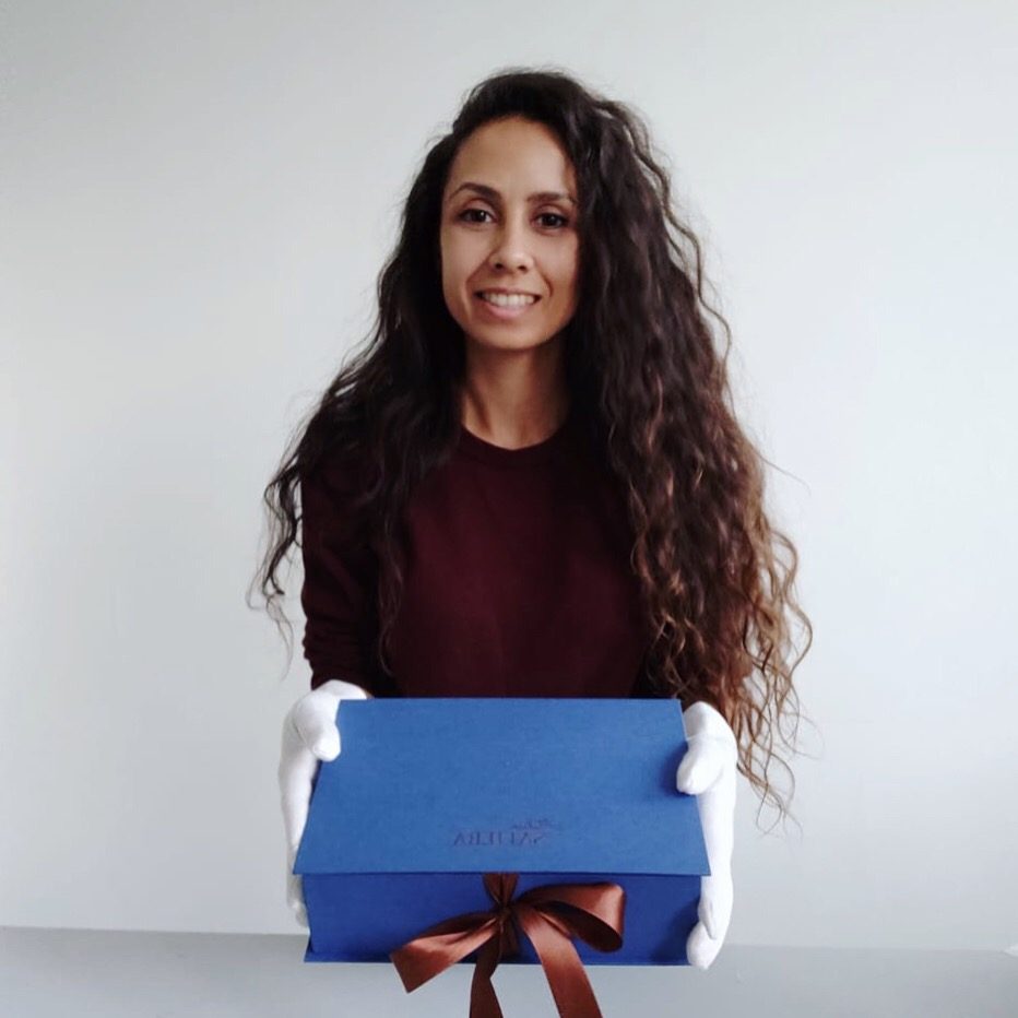 Safiera Pattipeilohij in handling gloves, holding a blue Safiera gift box tied with a copper ribbon