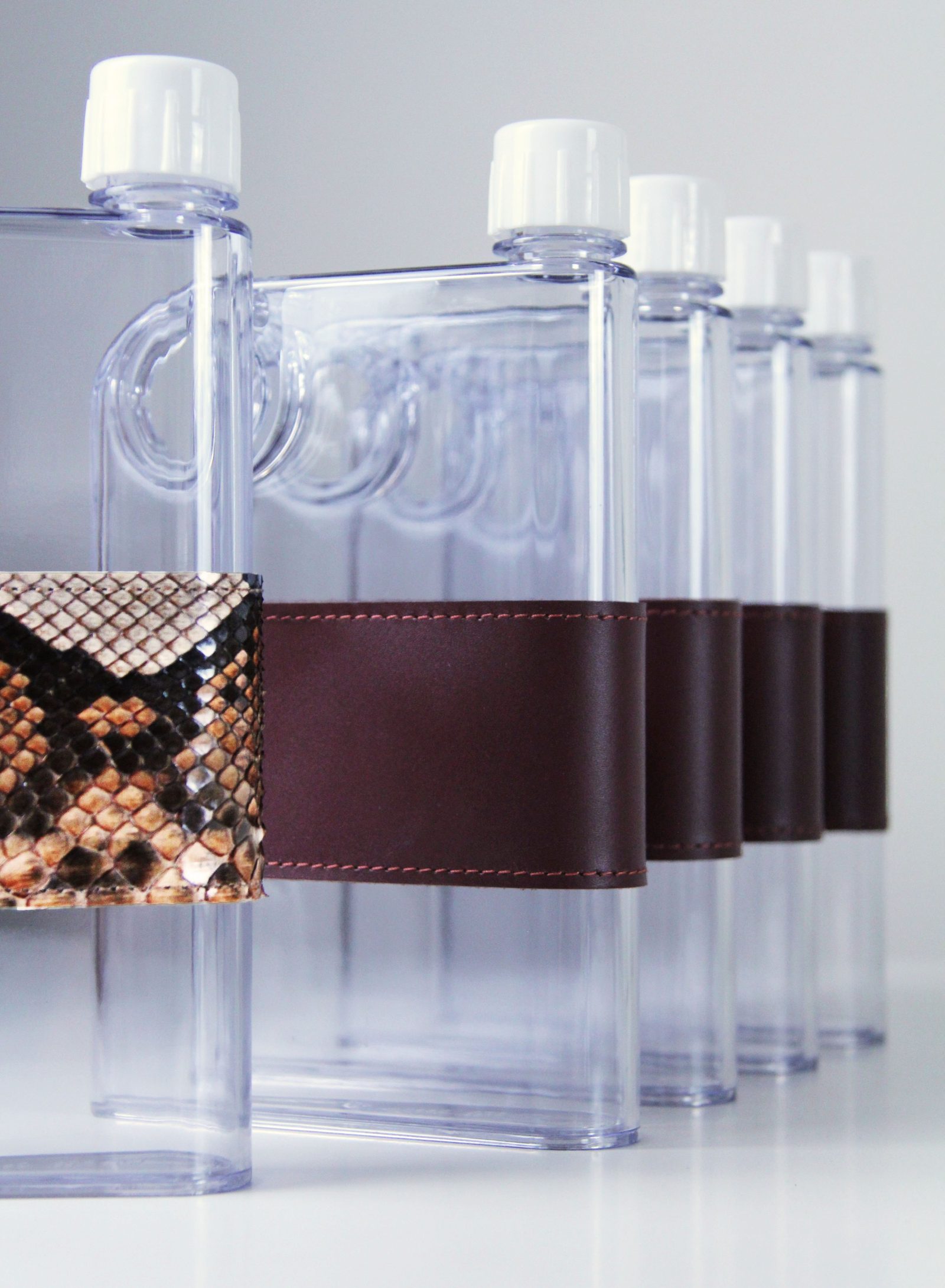 A row of flat Safiera water bottles, each wrapped in a leather band, the nearest in python-print leather