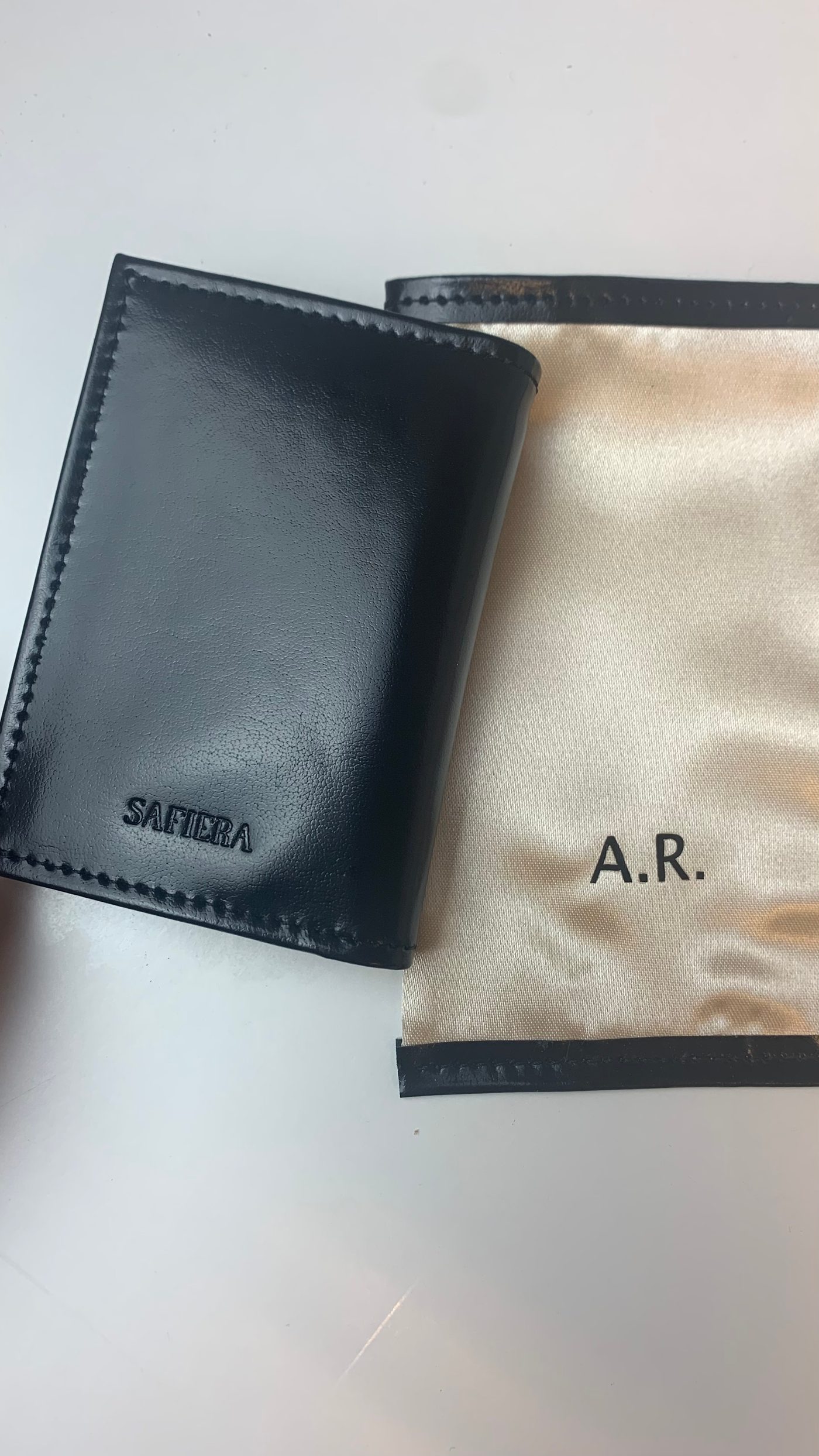 A black leather Safiera card wallet beside a satin pouch embossed with the initials A.R.