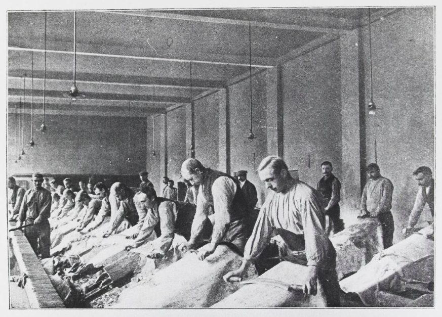 Rows of workers finishing leather by hand at long tables in a large hall