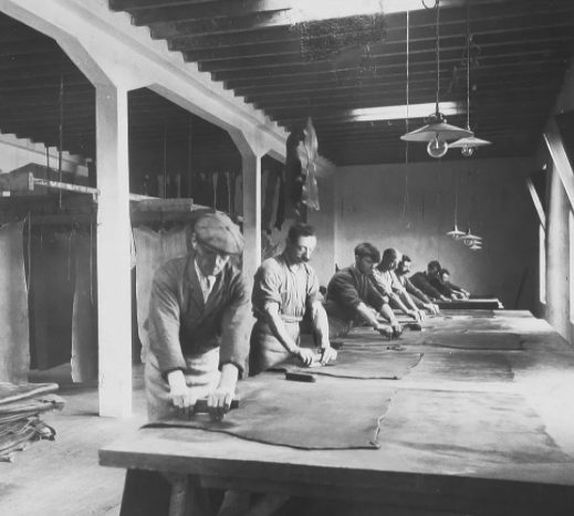Men working hides by hand at wooden tables under hanging lamps